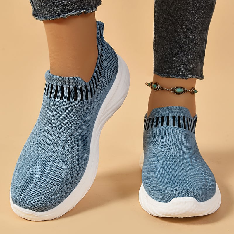Blaue Damen-Sneaker mit gestricktem Obermaterial und weißer Sohle, getragen mit Jeans und türkisfarbenem Fußkettchen. Bequeme, modische Schuhe.