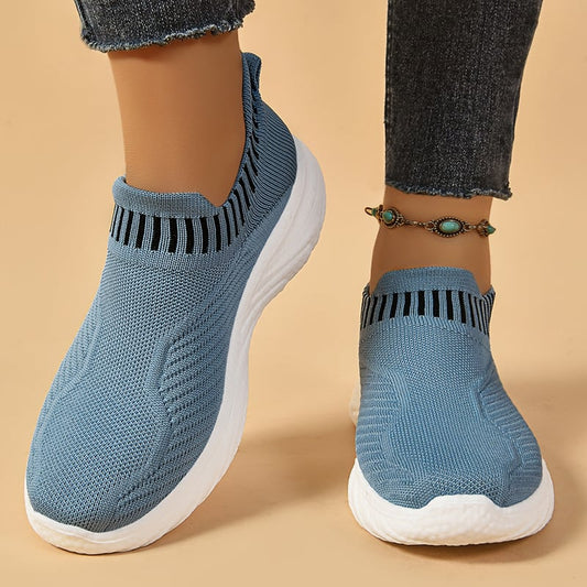 Blaue Damen-Sneaker mit gestricktem Obermaterial und weißer Sohle, getragen mit Jeans und türkisfarbenem Fußkettchen. Bequeme, modische Schuhe.