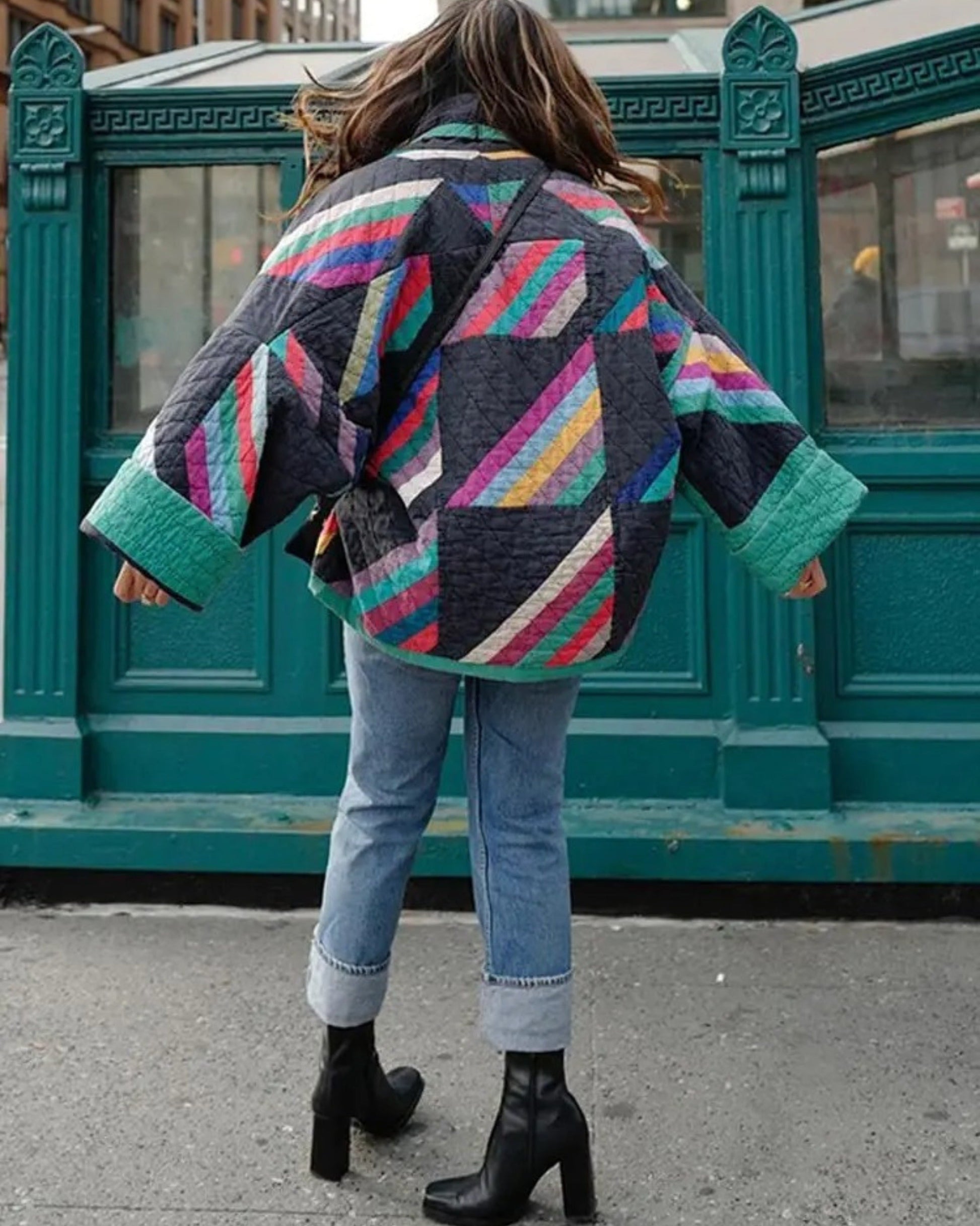 Frau in bunter Patchwork-Jacke und Jeans vor U-Bahn-Eingang, modische Herbstkleidung, Streetstyle, hohe Stiefel, urbaner Hintergrund.