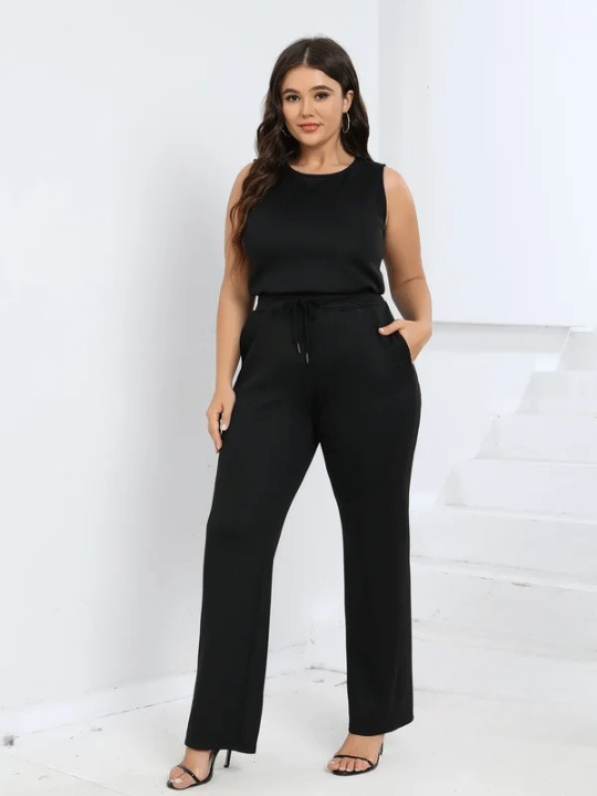Frau in eleganter schwarzer Hose und ärmellosem Top, steht vor weißem Hintergrund. Plus-Size-Mode, stilvoll, modern, selbstbewusst.