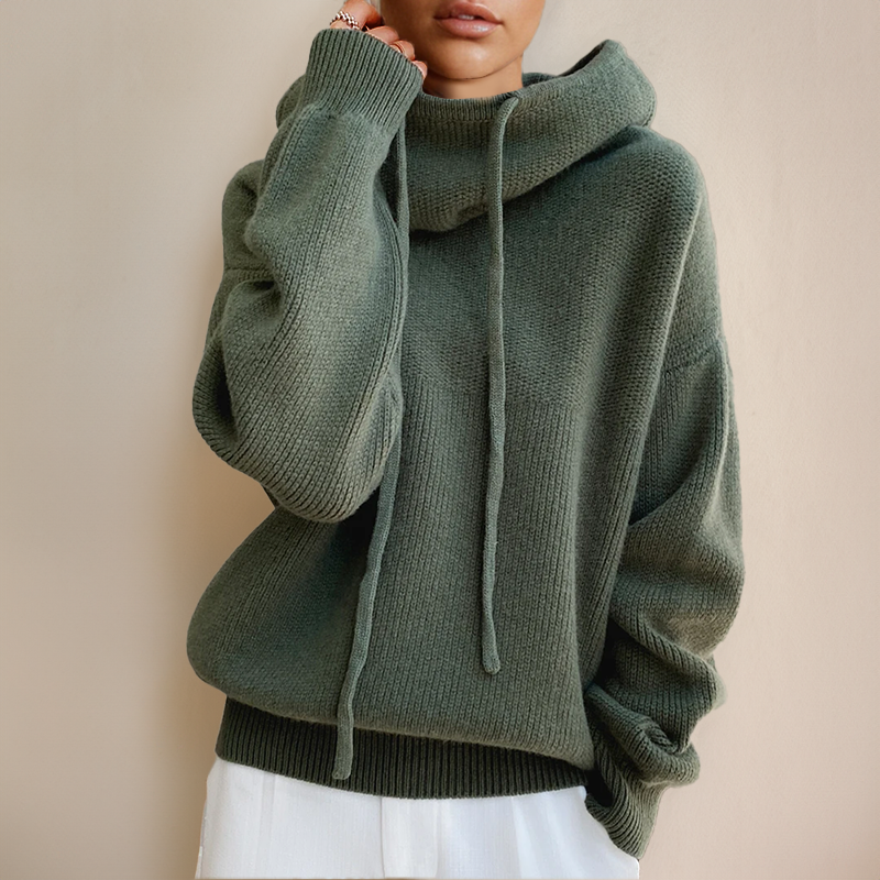 Frau trägt grünen Oversize-Pullover mit Kapuze und Kordeln, kombiniert mit weißer Hose. Modetrend Herbst/Winter, gemütlicher Stricklook.