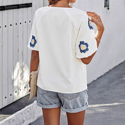 Frau in weißer Bluse mit blauen Blumenstickereien, Jeansshorts und Strohtasche, geht auf grauem Gehweg vor weißem Holztor.