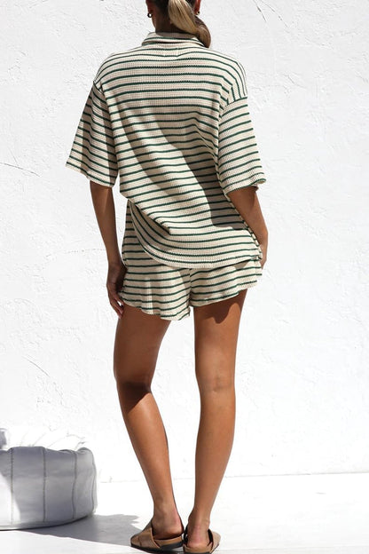 Frau in gestreiftem, lockerem Sommer-Set aus Shirt und Shorts, steht vor weißer Wand. Modetrend, lässiger Stil, bequeme Damenbekleidung.