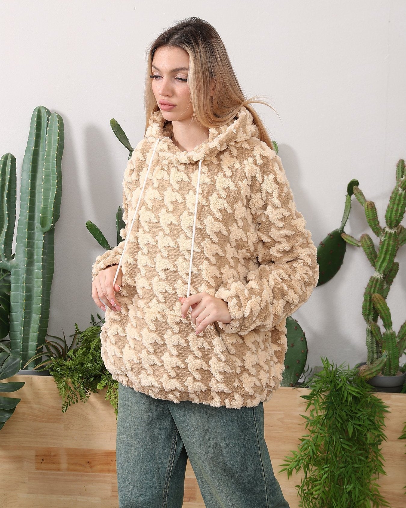 Frau in beigem Teddy-Pullover mit Kapuze, umgeben von Kakteen. Modetrend, gemütlicher Look, Herbstmode, lässiger Stil, Indoor-Pflanzen.