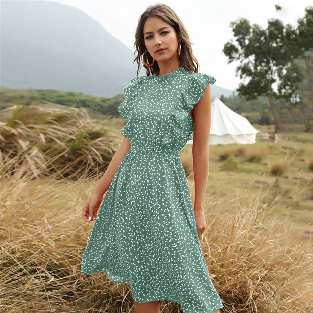 Frau in grünem, gepunktetem Kleid steht in natürlicher Landschaft mit Zelt im Hintergrund. Sommermode, Outdoor, Freizeitkleidung.