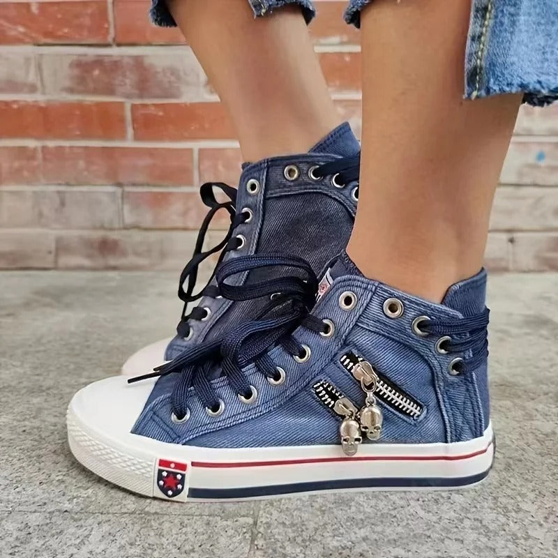 Blaue Jeans-Sneaker mit Reißverschluss und Totenkopf-Anhänger, weiße Gummisohle, schwarze Schnürsenkel, getragen von Person vor Ziegelwand.