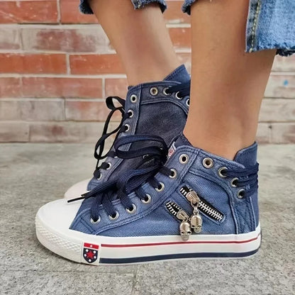 Blaue Jeans-Sneaker mit Reißverschluss und Totenkopf-Anhänger, weiße Gummisohle, schwarze Schnürsenkel, getragen von Person vor Ziegelwand.
