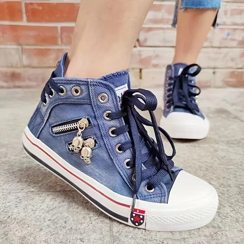 Blaue High-Top-Sneaker aus Denim mit weißen Sohlen, Zierreißverschluss und Totenkopf-Anhänger, getragen von einer Person auf Betonboden.
