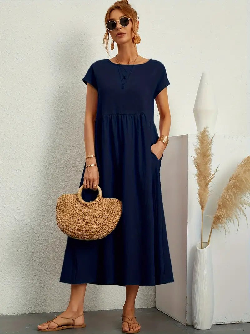 Frau in dunkelblauem Sommerkleid mit Strohtasche, Sonnenbrille und Sandalen, steht neben Vase mit Pampasgras. Elegante, lässige Mode.