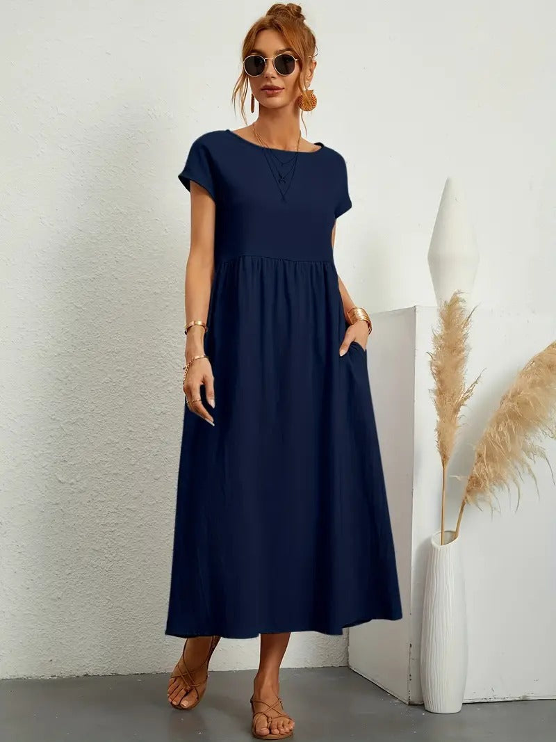 Frau in dunkelblauem Maxikleid, Sonnenbrille und Sandalen, steht neben Vase mit Pampasgras. Elegantes Sommeroutfit, lässiger Stil.