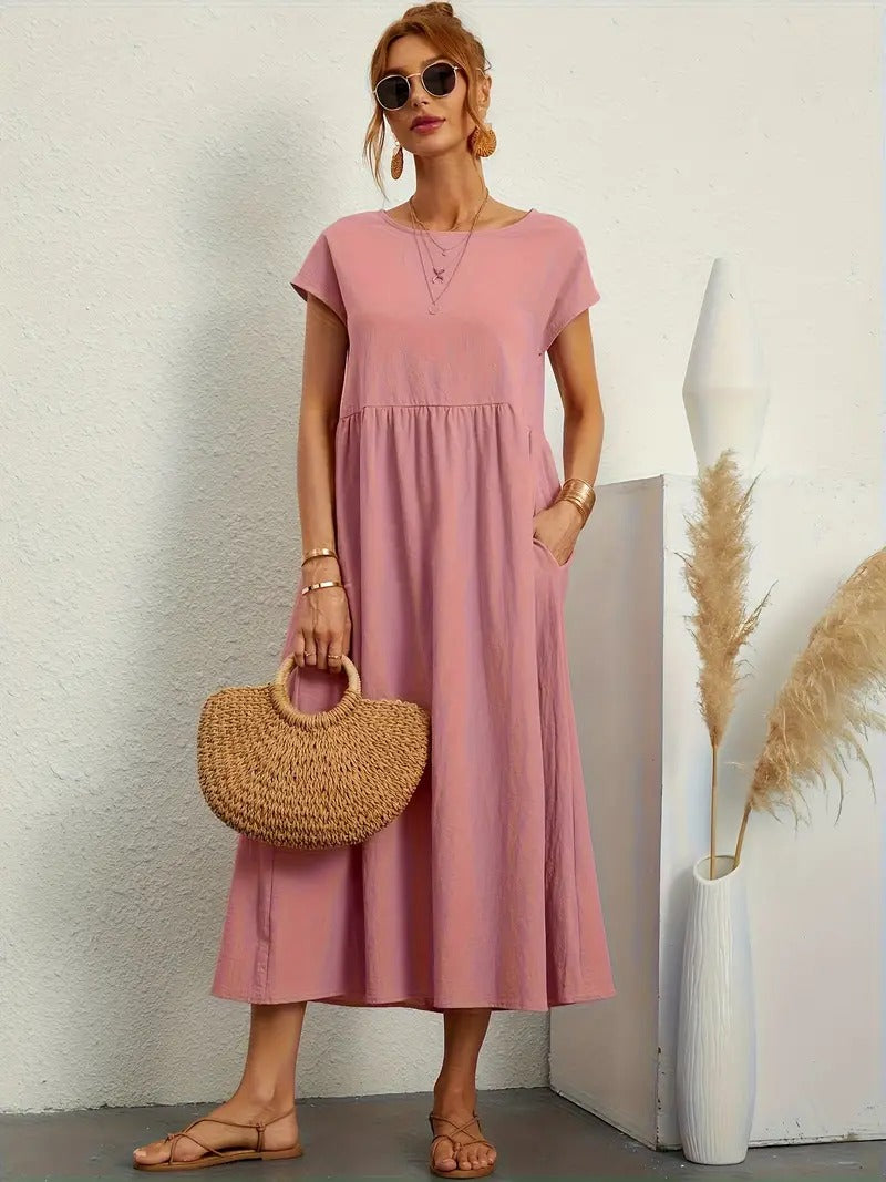 Frau in rosa Sommerkleid mit Strohtasche, Sonnenbrille und Sandalen, steht neben Vase mit Pampasgras. Elegante, lässige Mode für den Sommer.