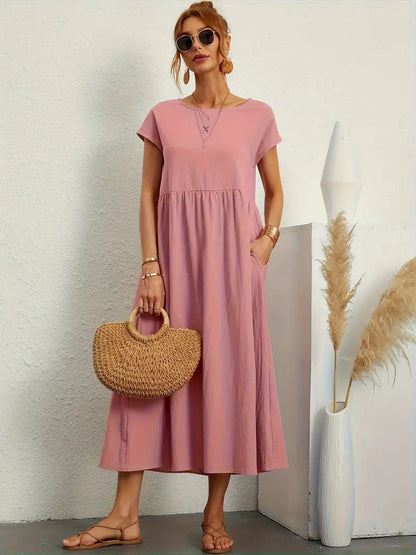 Frau in rosa Sommerkleid mit Strohtasche, Sonnenbrille und Sandalen, steht neben Vase mit Pampasgras. Elegante, lässige Mode für den Sommer.