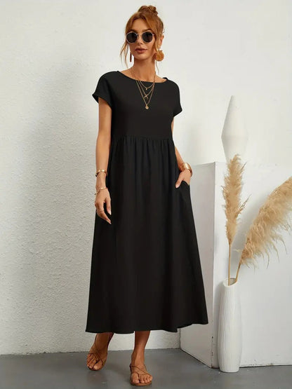 Frau in schwarzem Maxikleid mit kurzen Ärmeln, Sonnenbrille und Sandalen, steht neben Vase mit Pampasgras. Elegantes Sommeroutfit, lässiger Stil.