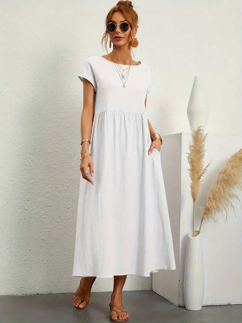 Frau in weißem Sommerkleid, Sonnenbrille, Sandalen, steht neben Vase mit Pampasgras. Elegante, lässige Mode, ideal für warme Tage.