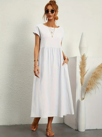Frau in weißem Sommerkleid, Sonnenbrille, Sandalen, steht neben Vase mit Pampasgras. Elegante, lässige Mode, ideal für warme Tage.