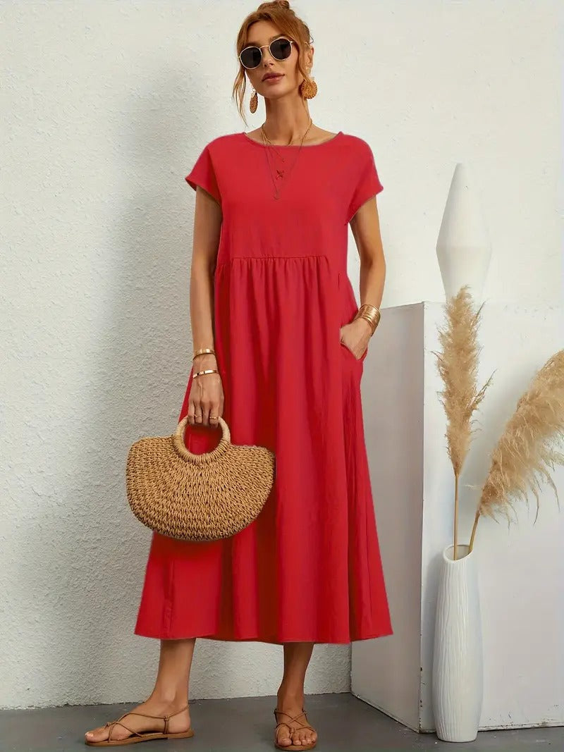 Frau in rotem Sommerkleid mit Strohtasche, Sonnenbrille und Sandalen. Steht vor weißer Wand mit Pampasgras in Vase. Elegante Sommermode.