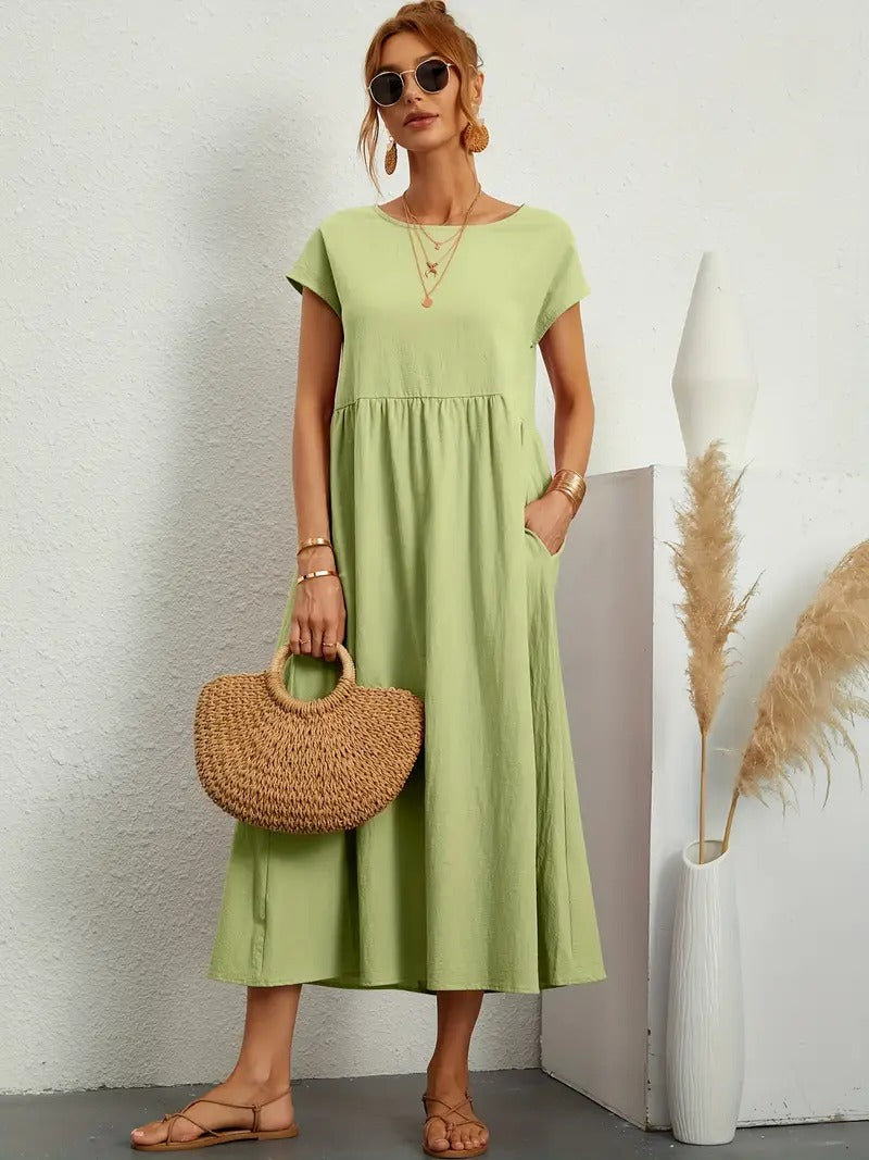 Frau in grünem Sommerkleid mit Strohtasche, Sonnenbrille und Sandalen. Steht vor weißer Wand mit Pampasgras in Vase. Elegante Sommermode.