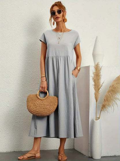 Frau in grauem Sommerkleid mit Strohtasche, Sonnenbrille und Sandalen vor weißer Wand. Mode, Damenbekleidung, Sommeroutfit, lässiger Stil.