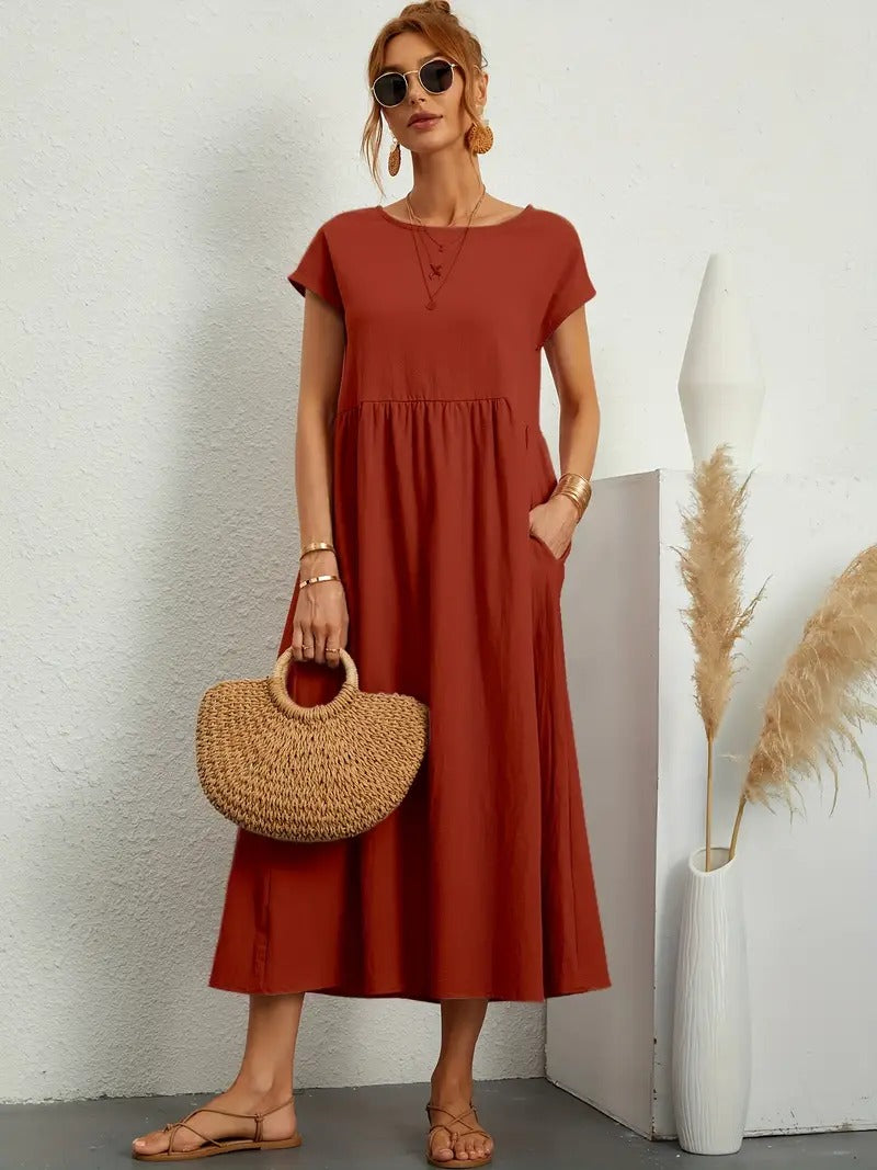Frau in rotem Sommerkleid mit Strohtasche, Sonnenbrille und Sandalen, steht vor weißer Wand mit Dekovase und Pampasgras. Elegante Sommermode.