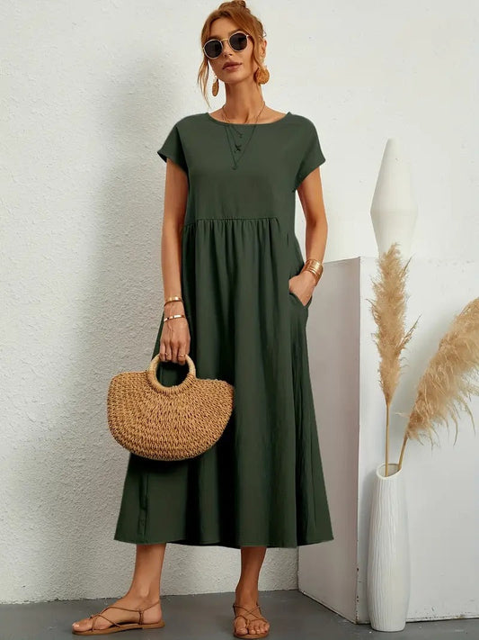 Frau in grünem Sommerkleid mit Strohtasche und Sandalen, steht vor weißer Wand. Mode, Damenbekleidung, Sommeroutfit, lässiger Stil.