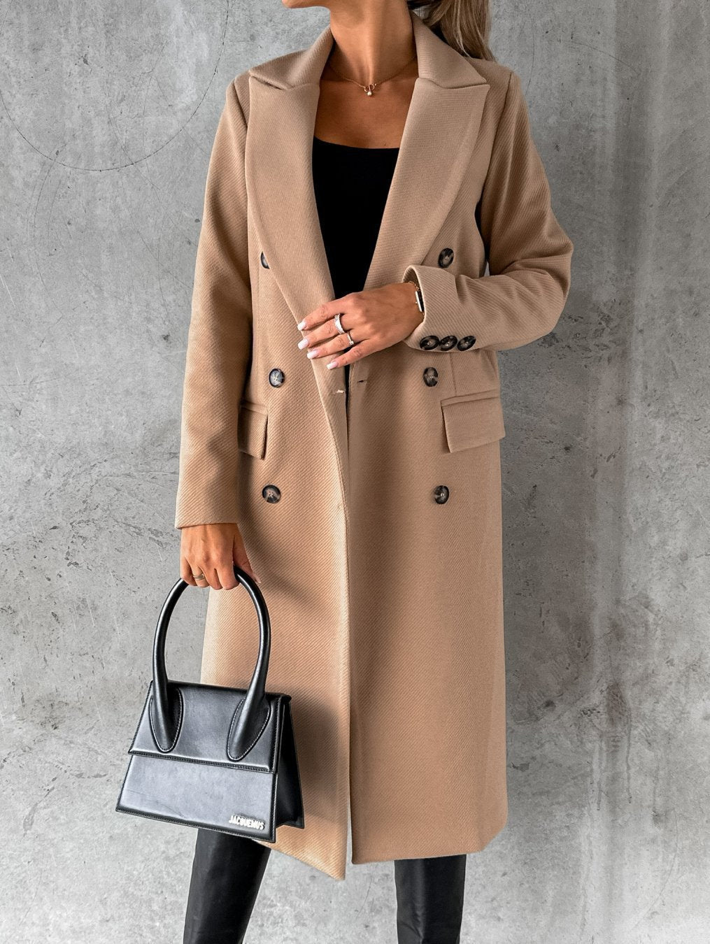 Eleganter beiger Damenmantel mit doppelter Knopfleiste, kombiniert mit schwarzer Handtasche und schwarzem Outfit, vor grauem Hintergrund.