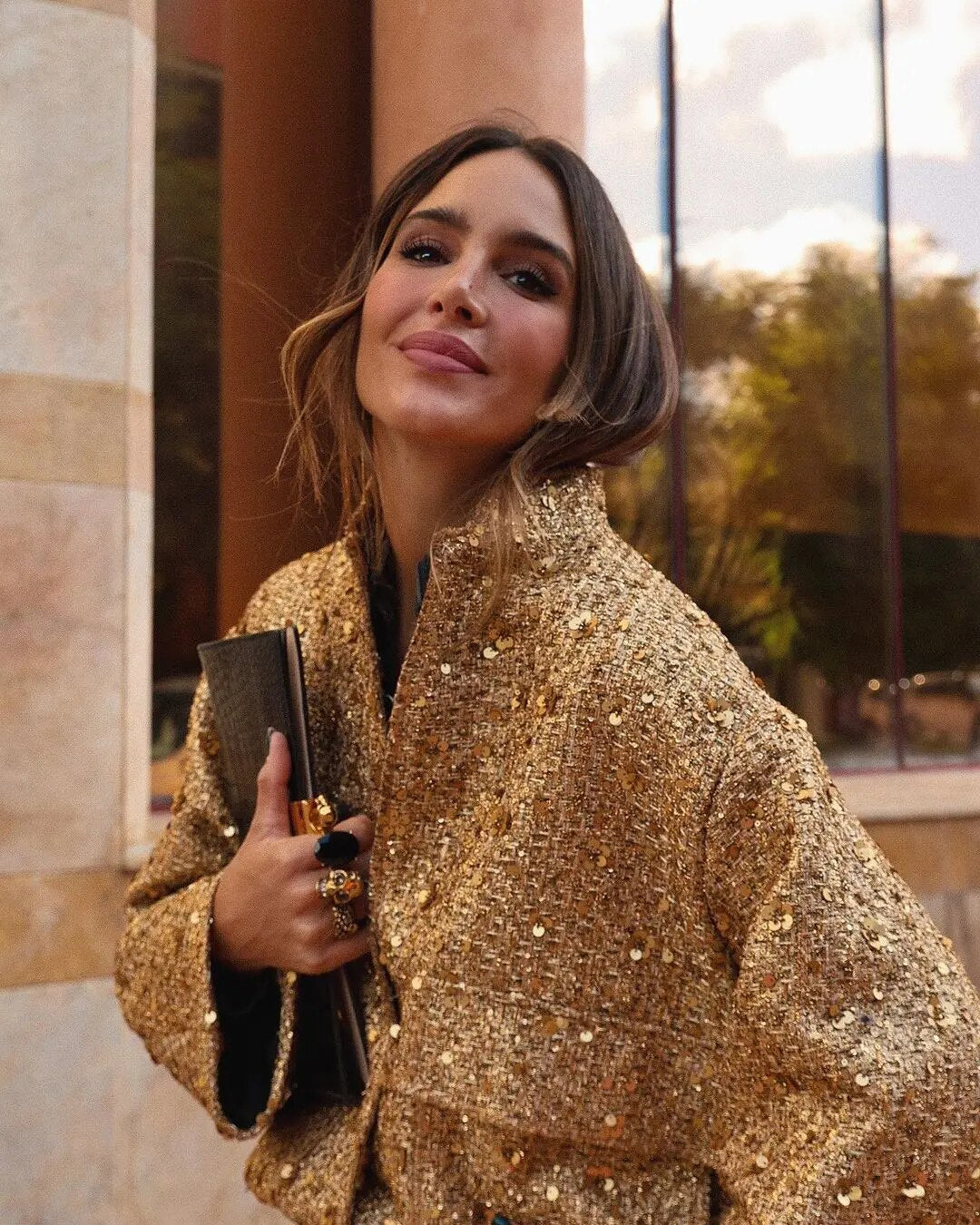 Frau in goldener Paillettenjacke, hält schwarze Clutch, lächelt vor modernem Gebäude. Elegante Mode, glamouröser Look, Herbststil.