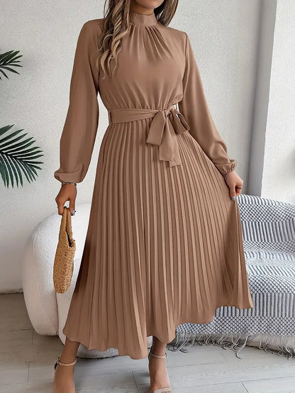Frau in elegantem, langem, braunem Plisseekleid mit Gürtel, hält eine Strohtasche. Perfekt für Herbstmode, stilvoll und modern.