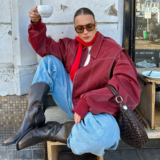 Frau in roter Jacke und blauer Jeans sitzt lässig mit Kaffeetasse, trägt Sonnenbrille und Stiefel. Streetstyle, Mode, urbane Kleidung.