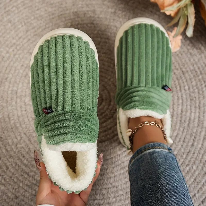 Grüne, plüschige Hausschuhe mit Rillenmuster auf grauem Teppich, getragen von Person in Jeans. Bequeme, warme Pantoffeln für Zuhause.