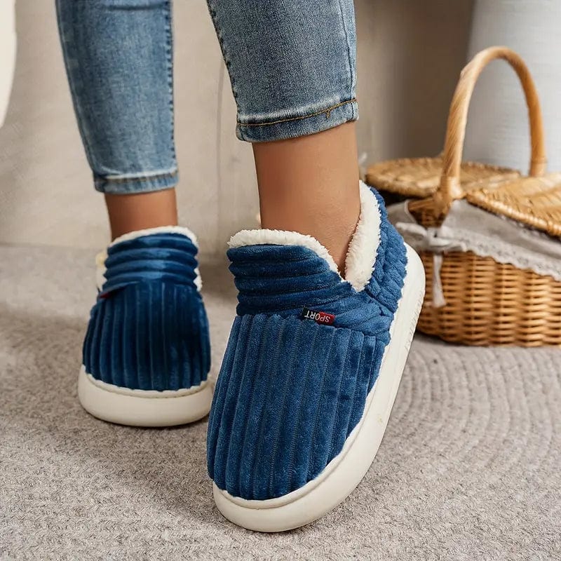 Blaue, gestreifte Hausschuhe mit weißem Futter an Füßen, getragen mit Jeans. Bequeme, warme Winterhausschuhe für Damen. Korb im Hintergrund.