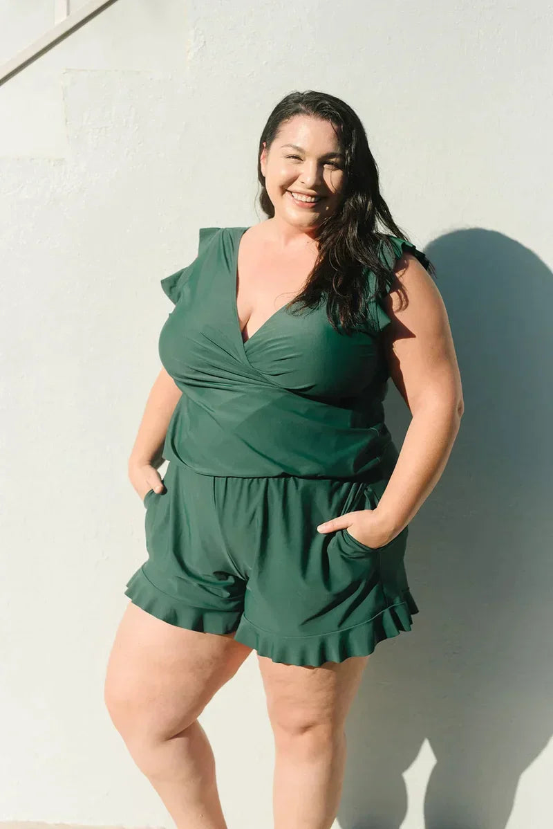 Frau in grünem Jumpsuit, lächelnd, vor weißer Wand. Plus-Size-Mode, Sommeroutfit, stilvoll, bequem, Freizeitkleidung.