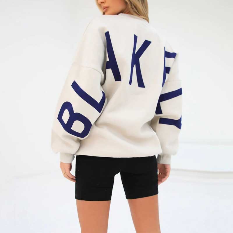 Frau in weißem Sweatshirt mit blauem "BLAKELY"-Schriftzug, schwarze Shorts, minimalistische Mode, Rückansicht, urbaner Stil.
