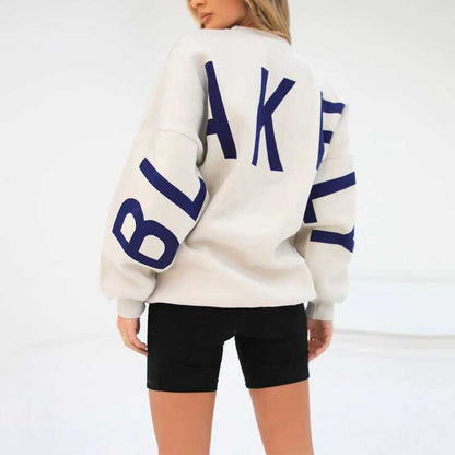 Frau in weißem Sweatshirt mit blauem "BLAKELY"-Schriftzug, schwarze Shorts, minimalistische Mode, Rückansicht, urbaner Stil.
