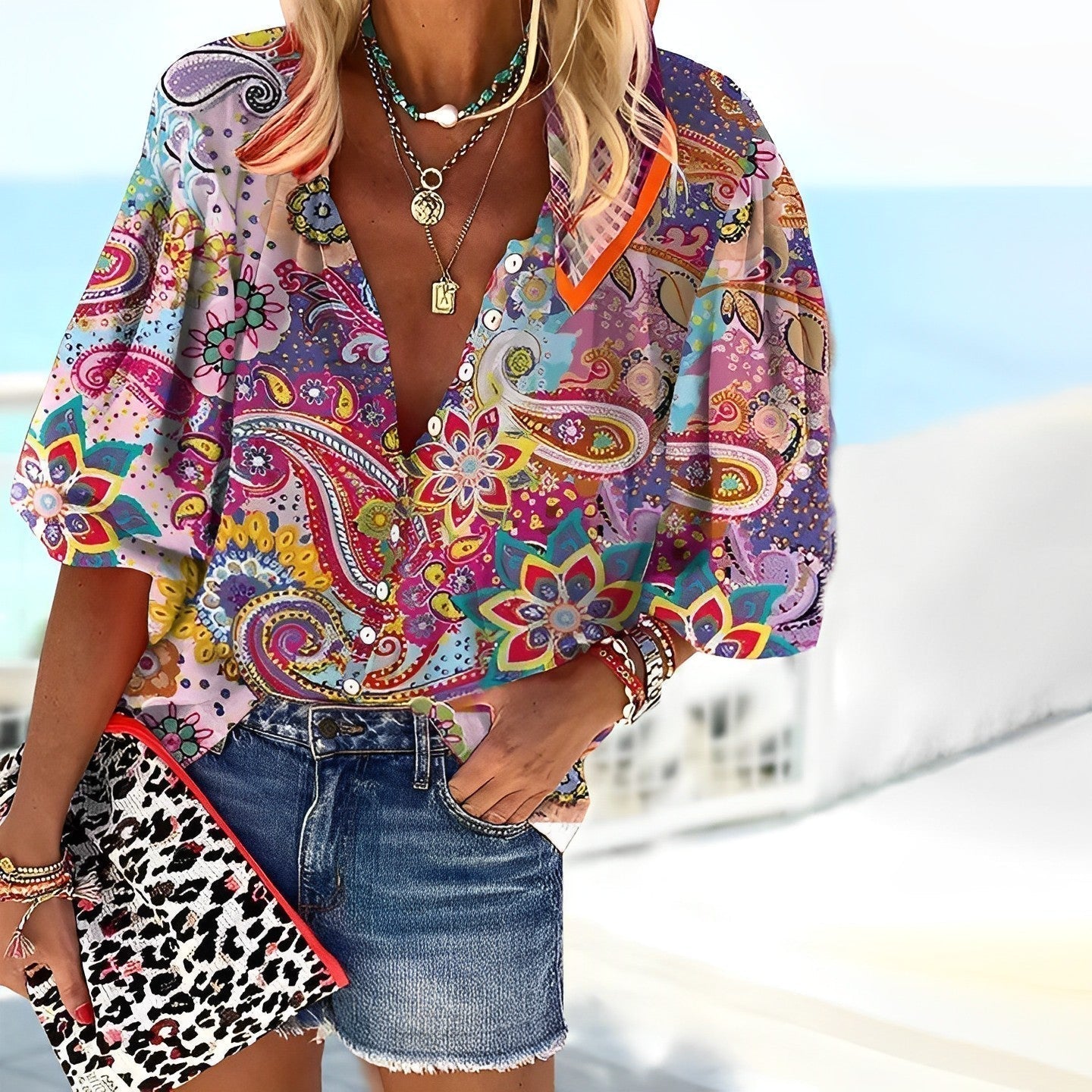 Frau in bunter Paisley-Bluse und Jeansshorts am Strand, hält eine Leoparden-Clutch. Sommermode, Boho-Stil, Strandoutfit, modische Accessoires.