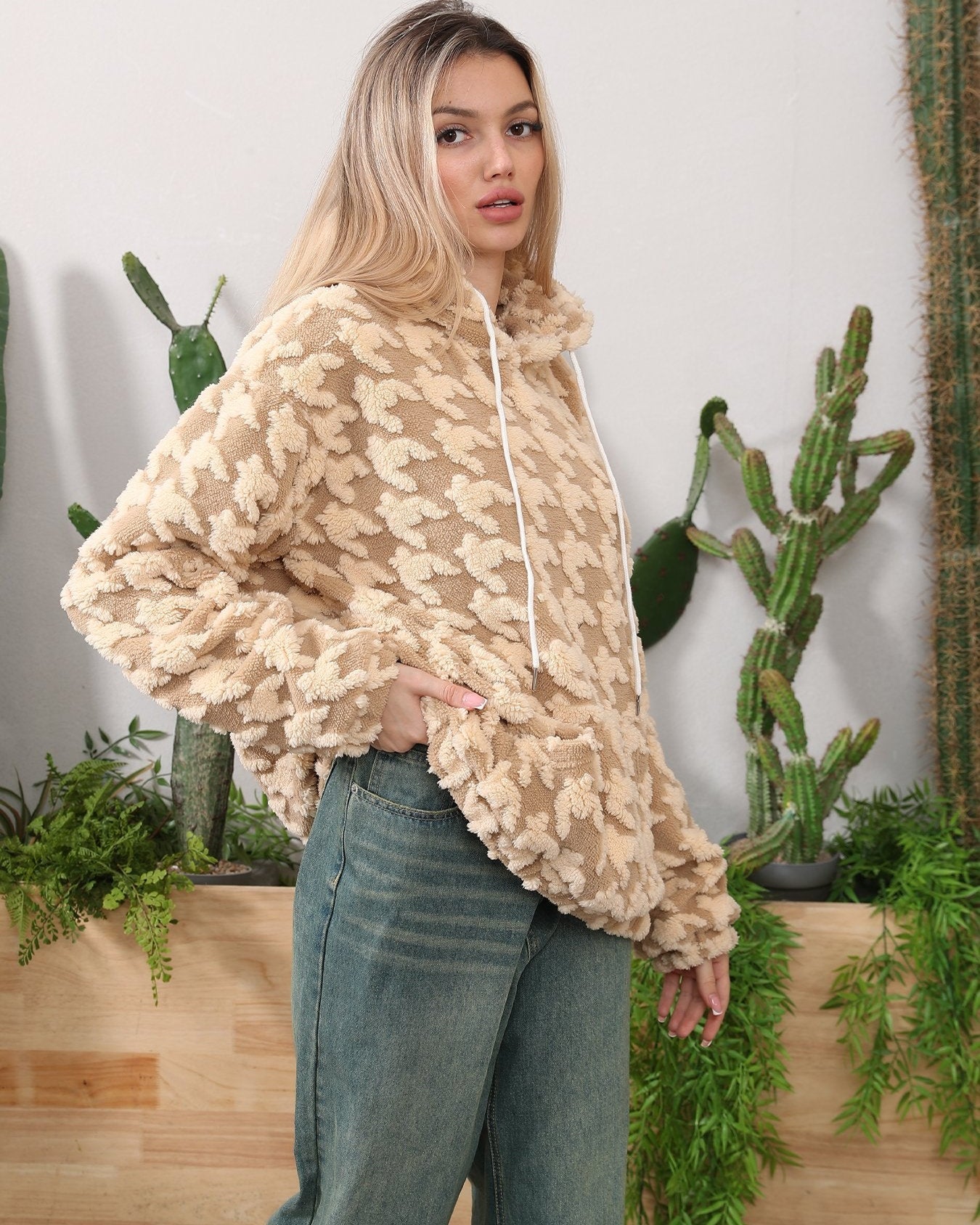 Frau in beigem, gemustertem Hoodie vor Kakteen, lässig gekleidet in Jeans. Indoor-Pflanzen, Mode, gemütlicher Look.