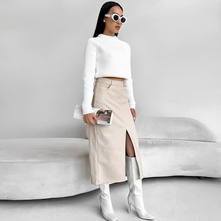 Frau in weißem Pullover und beigem Rock mit Schlitz, trägt weiße Stiefel und Sonnenbrille, steht vor grauem Sofa. Elegante Mode, minimalistischer Stil.