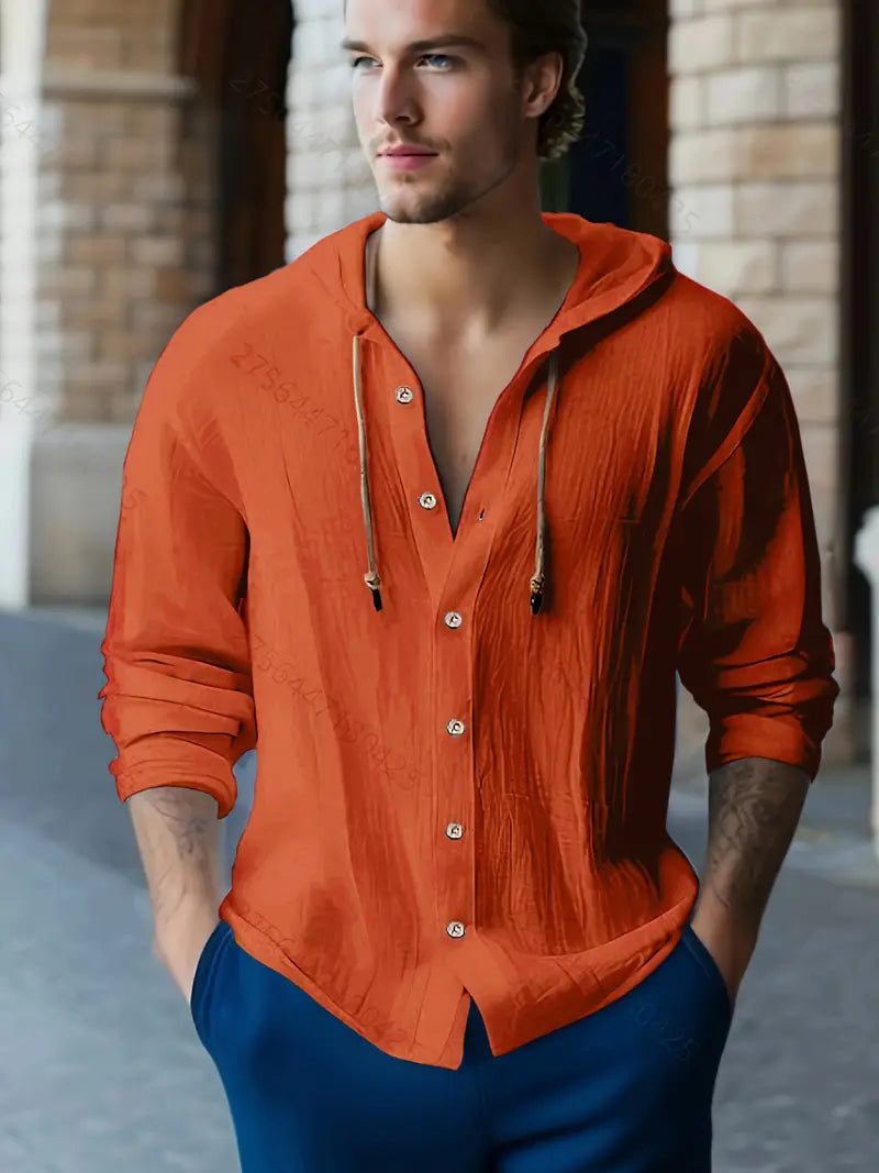 Junger Mann in orangefarbenem, langärmligem Hemd mit Kapuze und blauen Hosen, steht vor einem Gebäude mit Steinbögen. Mode, Herrenbekleidung, Freizeitlook.