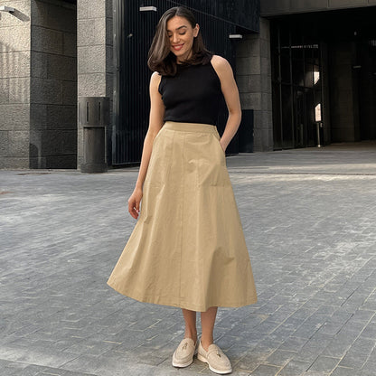 Frau in schwarzem Top und beigem Rock posiert lächelnd auf gepflastertem Weg vor modernem Gebäude. Elegante Mode, urbaner Stil.