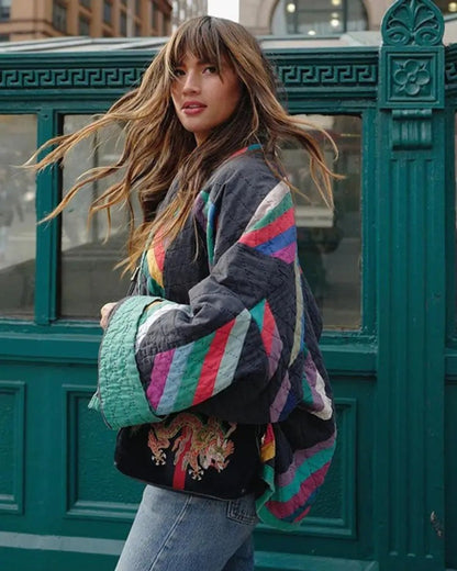 Frau mit buntem gestreiftem Mantel und Jeans vor grünem Gebäude. Modetrend, Streetstyle, Herbstmode, lässiger Look, Stadtleben.