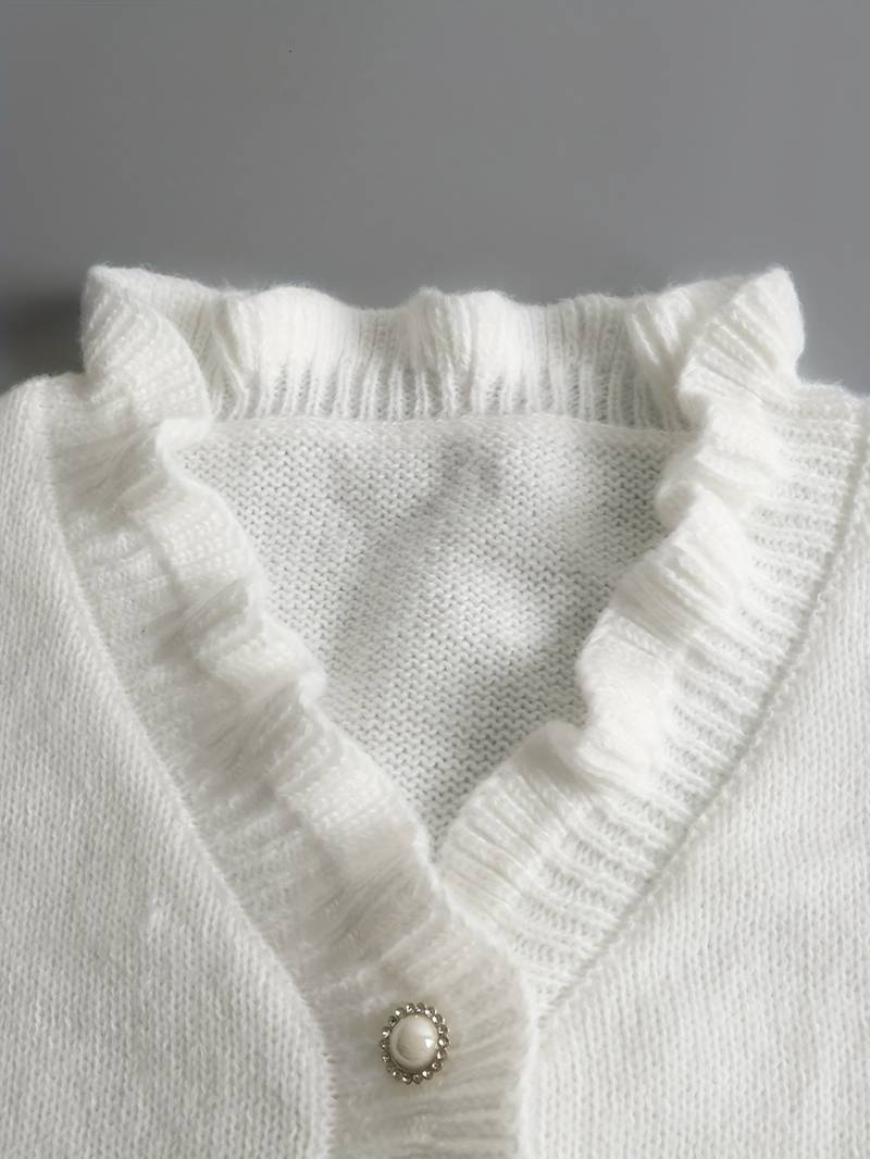 Weiße Strickjacke mit Rüschenkragen und Perlenknopf auf grauem Hintergrund. Elegante Damenmode, weicher Stoff, stilvolles Design.