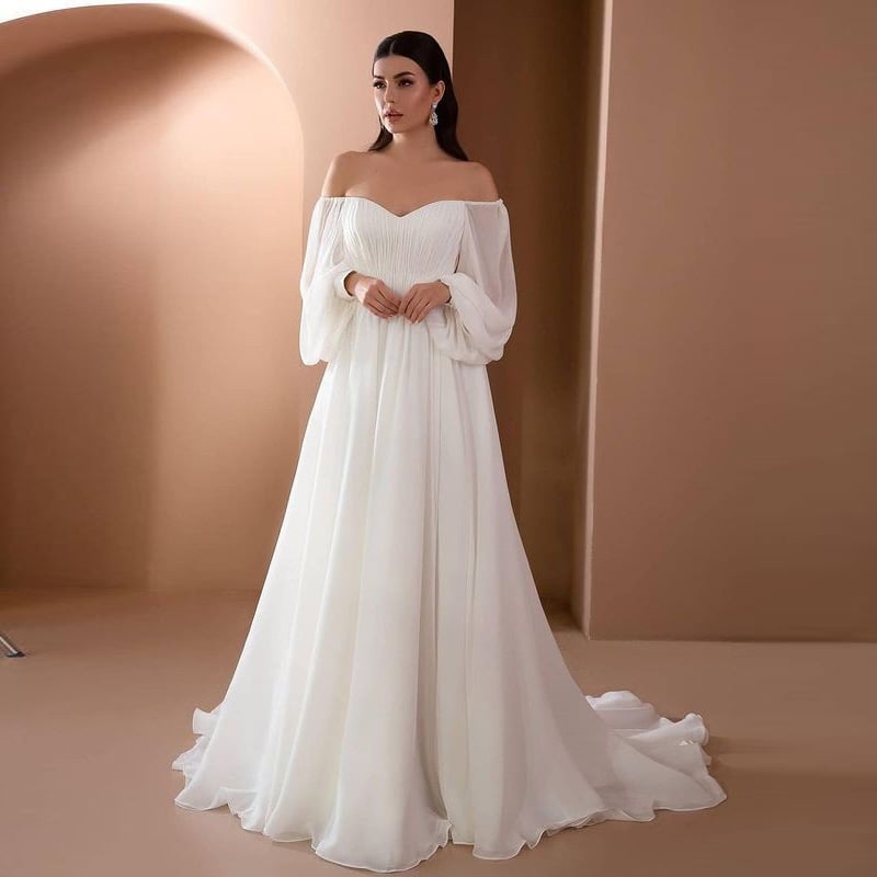 Braut in elegantem, schulterfreiem weißen Hochzeitskleid mit langen Ärmeln vor beigem Hintergrund. Perfektes Design für moderne Hochzeiten.