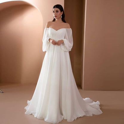 Braut in elegantem, schulterfreiem weißen Hochzeitskleid mit langen Ärmeln vor beigem Hintergrund. Perfektes Design für moderne Hochzeiten.