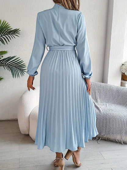 Frau in hellblauem, langem, plissiertem Kleid, steht in modernem Wohnzimmer. Elegantes Modeoutfit, Rückansicht, stilvolle Inneneinrichtung.