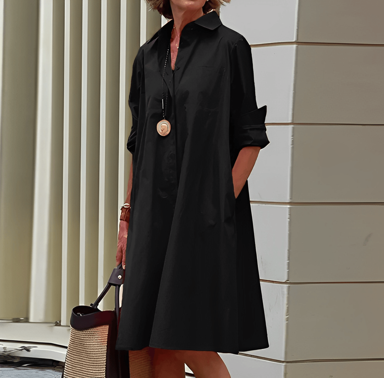 Frau in schwarzem Hemdkleid mit langen Ärmeln, trägt eine lange Halskette. Steht vor moderner Architektur. Elegantes, minimalistisches Modeoutfit.