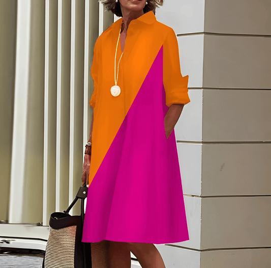 Frau in einem farbenfrohen Kleid mit orange-pinkem Design, steht vor moderner Architektur, trägt eine Halskette und hält eine Strohtasche.