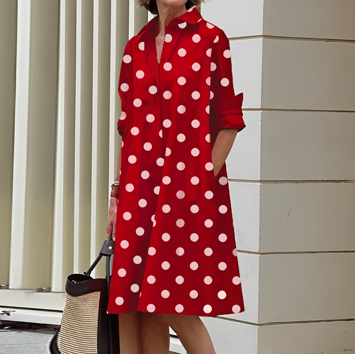 Frau in rotem, gepunktetem Kleid mit langen Ärmeln, hält eine Strohtasche. Stilvolle Mode, Sommerkleid, Polka-Dots, urbane Umgebung.