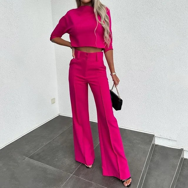 Frau in pinkem Hosenanzug mit weitem Bein und kurzem Oberteil, steht auf grauen Fliesen, hält eine schwarze Handtasche. Elegante Damenmode.