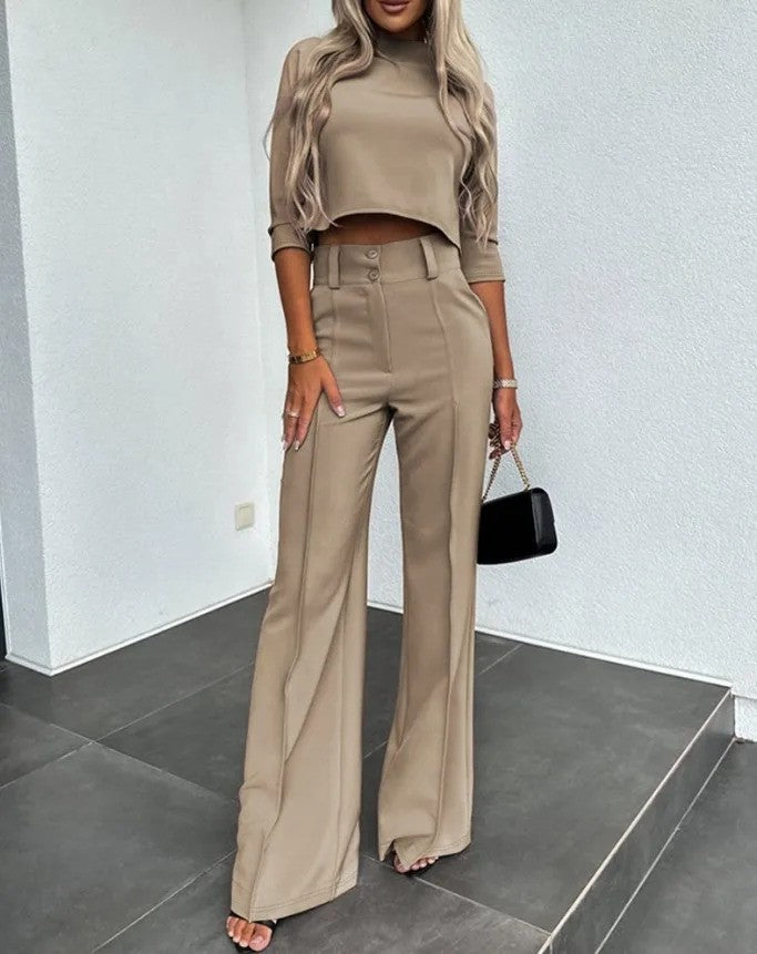 Frau in beigem, elegantem Zweiteiler mit weitem Bein, steht auf grauem Boden, hält schwarze Handtasche. Modetrend, Damenmode, stilvoller Look.