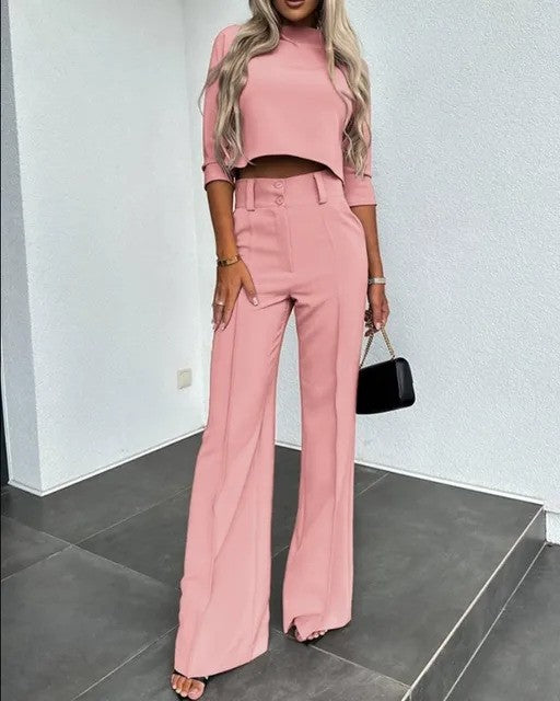 Frau in rosa Anzug mit weitem Bein und Crop-Top, steht auf grauem Boden, hält schwarze Handtasche. Elegante Damenmode, trendiger Look.