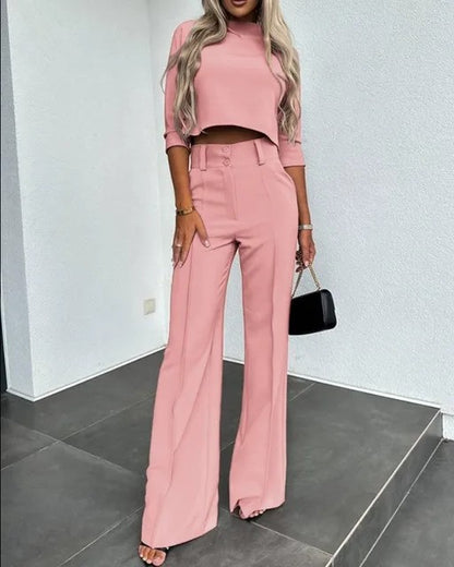 Frau in rosa Anzug mit weitem Bein und Crop-Top, steht auf grauem Boden, hält schwarze Handtasche. Elegante Damenmode, trendiger Look.
